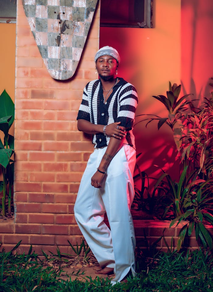 Fashionable man in striped top and white pants leaning against a brick wall outdoors.